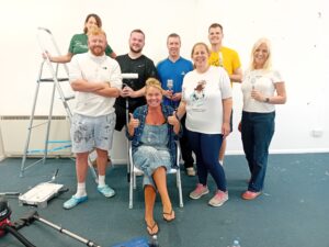 Seven volunteers from Lloyds holding paintbrushes and rollers with Caring Together Charity Shop Manager Belinda Jones sat in the middle of the group with thumbs up.